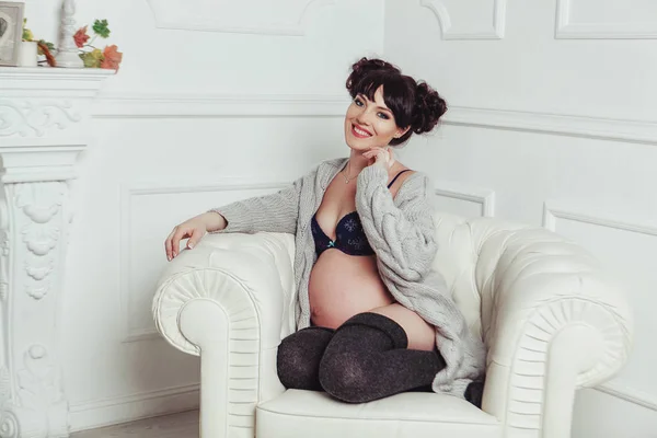 Sorrindo mulher grávida sentada em um sofá no estúdio interior branco — Fotografia de Stock