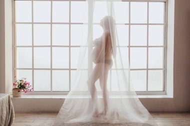 Şifon perde ile örtülü pencerenin önünde duran güzel genç anne.