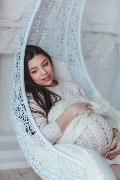 Junge schwangere Frau in weißem Strickpullover, die zu Hause im Sessel sitzt und ihren Bauch berührt. Konzept Schwangerschaft und Erwartung — Stockfoto