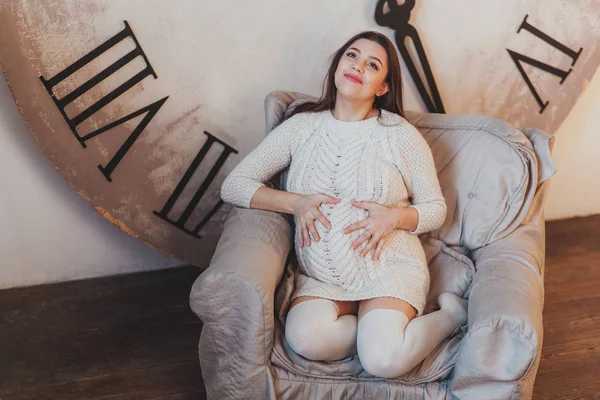 Pensive mulher grávida sonhando com criança sentada em poltrona com enorme relógio atrás — Fotografia de Stock