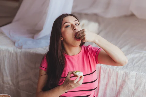 Atractiva mujer embarazada de pelo negro comiendo pastel con las manos. Primer plano estudio retrato en el dormitorio — Foto de Stock