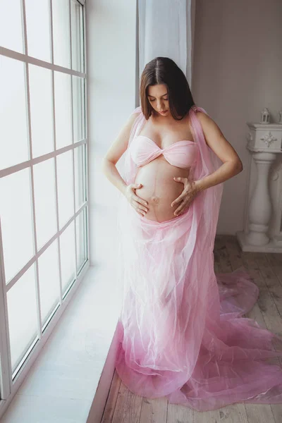 Πορτραίτο του διαγωνισμού ομορφιάς έγκυος νεαρή γυναίκα στέκεται μπροστά από το παράθυρο και να αγγίζουν την κοιλιά της στο σπίτι. Μινιμαλιστικό εσωτερικό πατάρι — Φωτογραφία Αρχείου