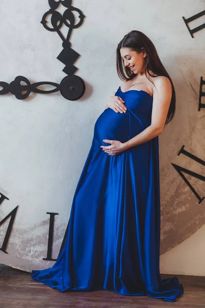 Uma mulher grávida em um belo vestido longo azul em pé na frente de um relógio enorme — Fotografia de Stock