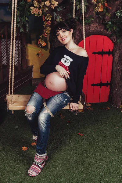 Menina grávida sentado em um balanço com madeira e porta vermelha no fundo — Fotografia de Stock