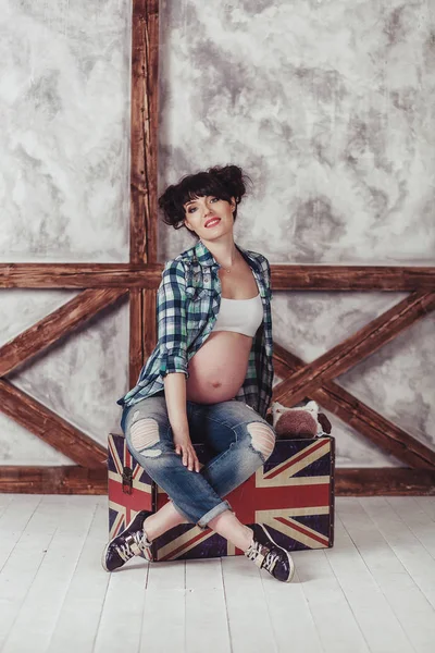 Uma jovem grávida sentada em uma mala velha contra a parede com tábuas de madeira . — Fotografia de Stock