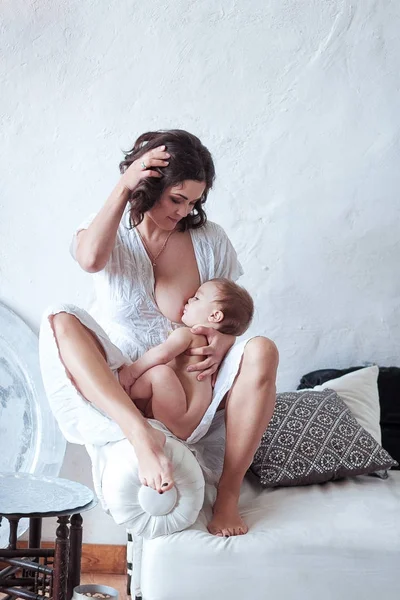 Uma jovem mulher amamentando bebê sentado no sofá com fundo de parede rústico branco — Fotografia de Stock