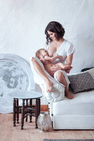 Uma jovem mulher amamentando bebê sentado no sofá com fundo de parede rústico branco — Fotografia de Stock