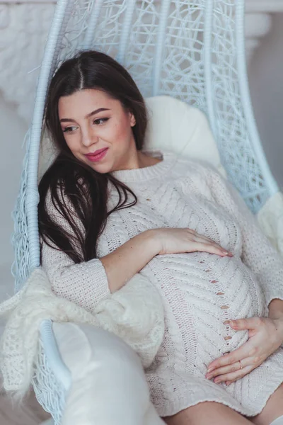 Junge schwangere Frau in weißem Strickpullover, die zu Hause im Sessel sitzt und ihren Bauch berührt. Konzept Schwangerschaft und Erwartung — Stockfoto