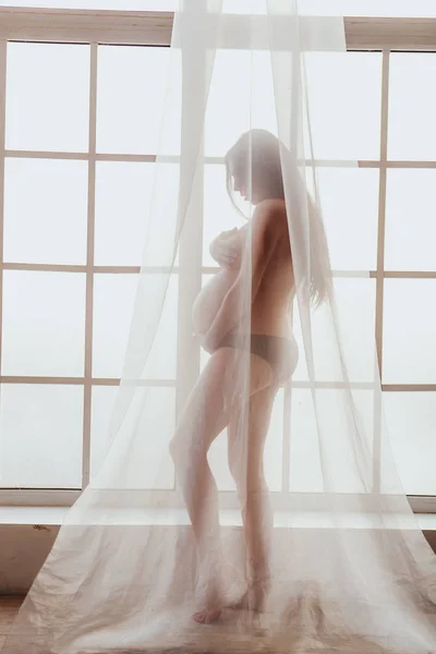 Bela jovem mãe de pé na frente da janela coberta com cortina de chiffon . — Fotografia de Stock