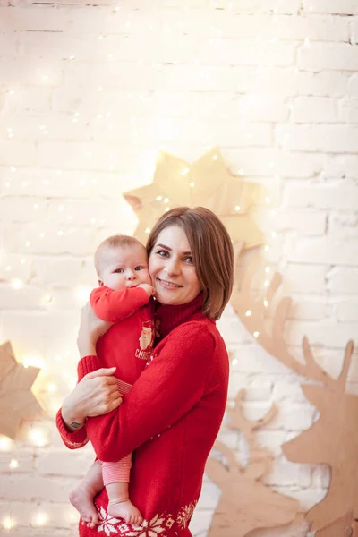 Uma jovem mãe e bebê vestida para o evento de Natal posando na frente da parede de tijolo branco com luz guirlanda e decoração de veados de madeira — Fotografia de Stock