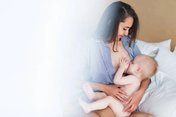 Μια νεαρή μητέρα κρατά το παιδί της το μωρό. Νοσηλευτική το μωρό μαμά. Γυναίκα θηλάζει νεογέννητο αγόρι σε μια λευκή κρεβατοκάμαρα. Λευκό επίδραση — Φωτογραφία Αρχείου