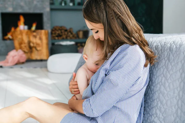 Bebé a amamentar. Jovem mãe segurando seu filho recém-nascido no quarto com lareira no fundo . — Fotografia de Stock