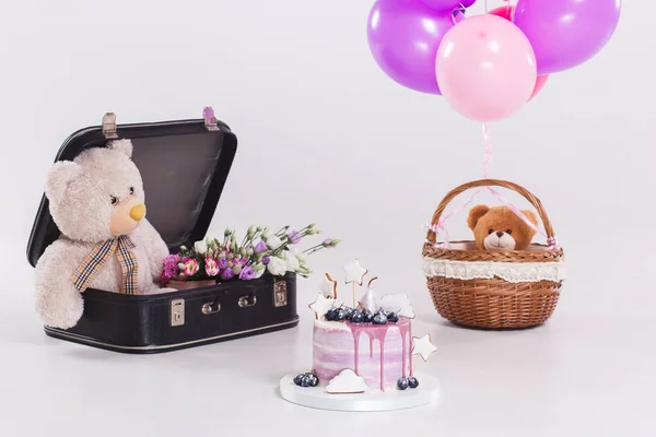 Modern round lilac birthday cake, balloons, teddy bear in vintage suitcase isolated.
