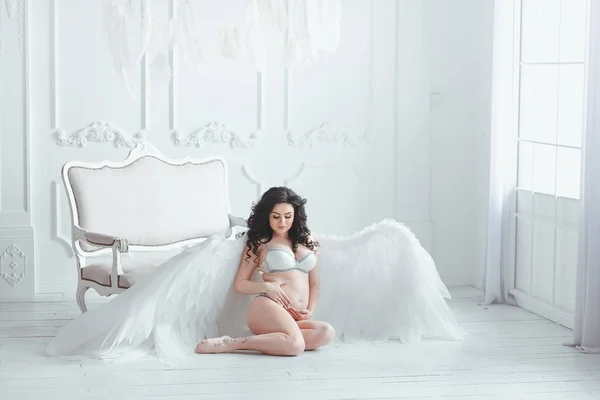 Menina grávida bonita com grandes asas de anjo no estúdio branco. Sentado no chão perto do sofá vintage — Fotografia de Stock