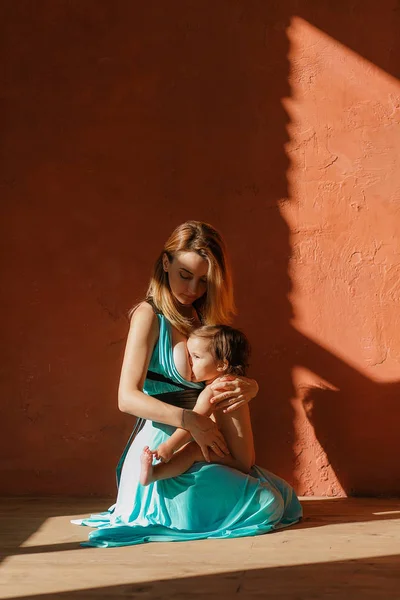 Μια νεαρή γυναίκα που θηλάζετε ένα μωρό που κάθονται στο πάτωμα, κεραμιδί τοίχο στο φόντο. Απομονωμένο στην ηλιαχτίδες — Φωτογραφία Αρχείου
