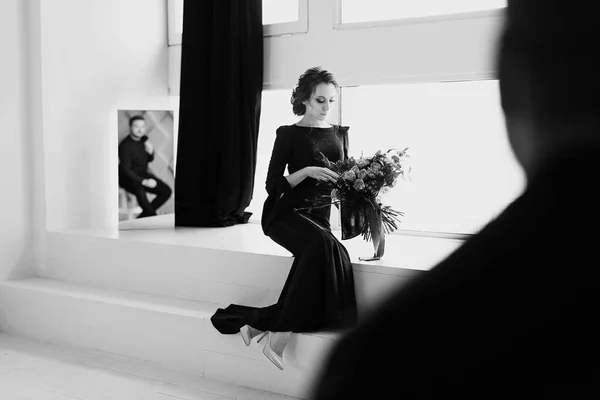 La mariée en robe noire. Réfléchissant dans le miroir. Studio blanc avec fenêtres. Plan noir et blanc avec silhouette floue du marié — Photo