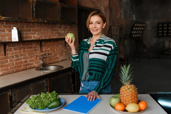 Hermosa mujer joven sana comiendo manzana en su moderna cocina loft. Entrenador. Come bien para mantener tu barriga apretada. —  Fotos de Stock