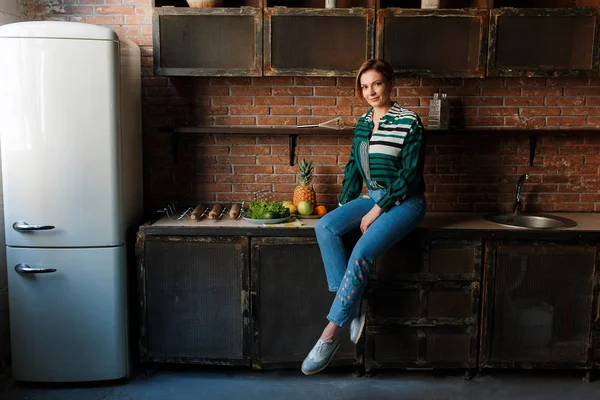 Hermosa mujer joven corte de pelo corto sentado en la mesa de la cocina. Fondo de los alrededores de la cocina estilo loft —  Fotos de Stock