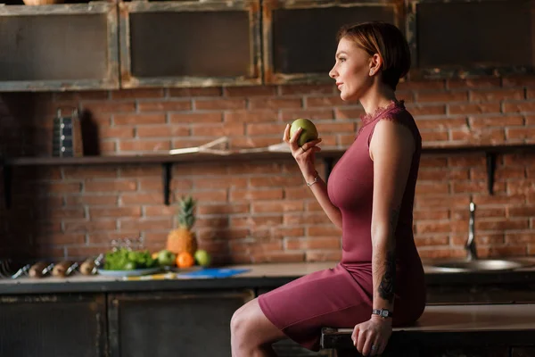 Szép nő ül a konyhaasztalnál, tart alma. Mosolygó fiatal nő modell pózol a téglafal háttér, írja a napló — Stock Fotó