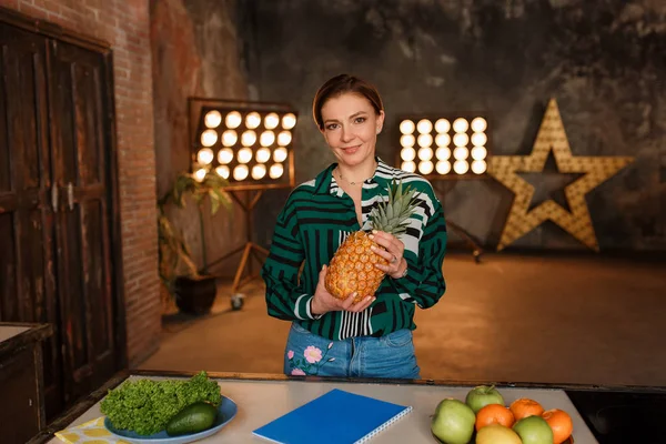 Fitness, home and diet concept. Young woman holding pineapple standing at kitchen table with retro bulb light on background