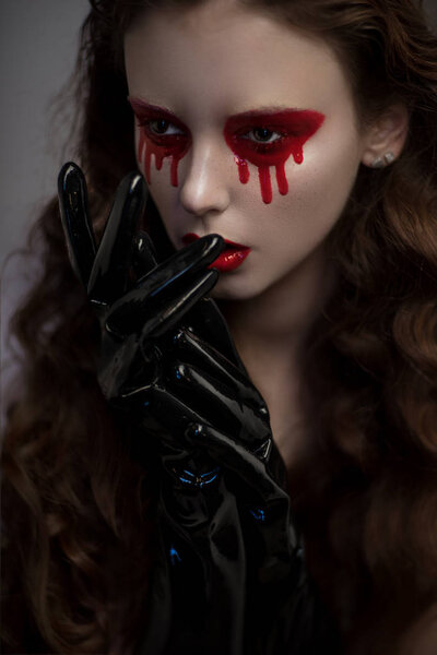 Portrait of beautiful woman with bloody eyes and latex black gloves on grey background