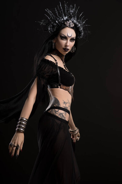 Beautiful woman in black gothic suit and horns crown posing at camera against black background