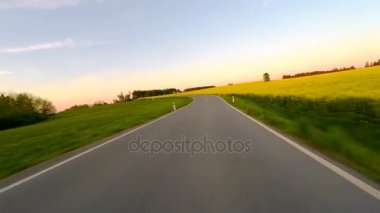 Bahar kırsal kırsal yol güneşli bir günde akşam araba araba. Manzara çayır ve tecavüz alan. Kamera önünde araç. Gün batımı ile mavi gökyüzü