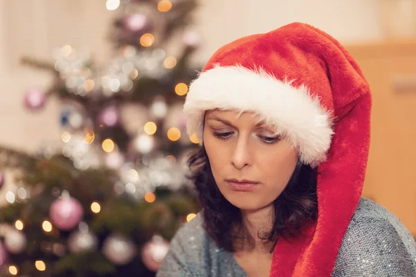 Vackra medelålders kvinna i santa hatt sitter nära granen漂亮的中年女子在圣诞树旁的圣诞老人的帽子 — 图库照片