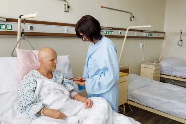 化学療法後の髪のない女性患者は 病院のベッドに横たわって悲しみと憂うつな心配感 友人が彼女の携帯電話に写真を見せに来て — ストック写真
