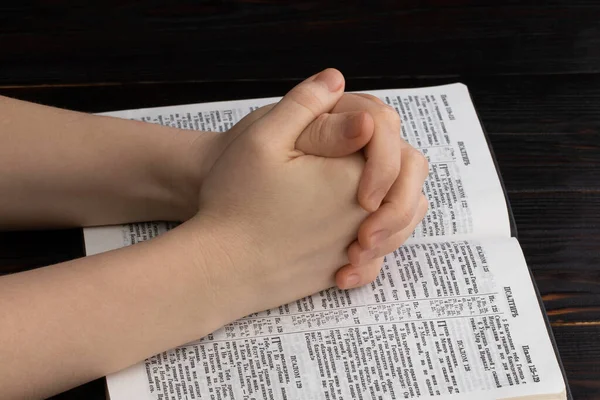 hands praying with a bible in a dark over wooden table.