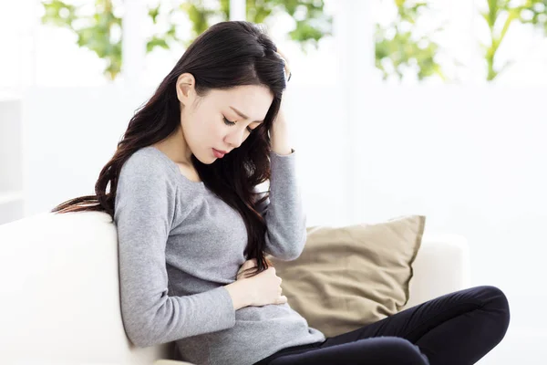 Mujer joven sentada en el sofá y con dolor de estómago —  Fotos de Stock
