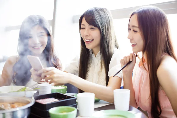 Fiatal nők csoport étkezési forró fazékba, és figyelembe képet a telefon — Stock Fotó