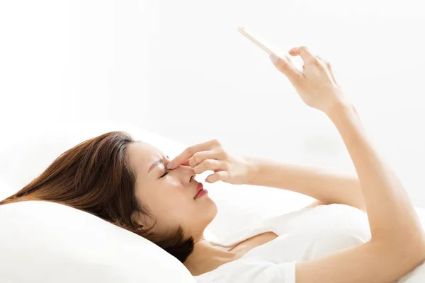 Junge Frau sucht Smartphone mit müden Augen im Bett — Stockfoto