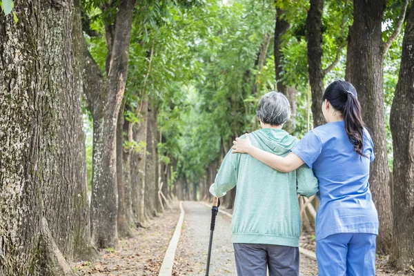 Perawat tersenyum membantu wanita tua berjalan-jalan di sekitar taman — Stok Foto