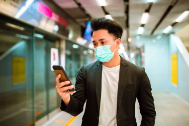 Yüz maskesi takan genç bir iş adamı cep telefonunu izliyor ve treni bekliyor. 