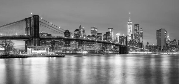 Panoramic view of Brooklyn Bridge and Lower Manhattan skyline in New York City at night with city illumination, USA. Black and white toned