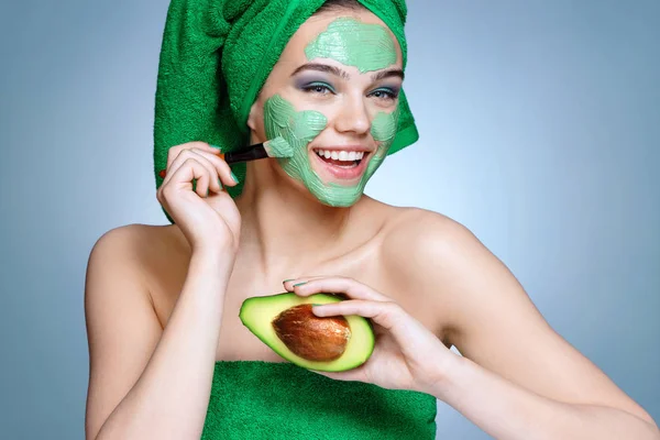 Happy girl receiving cosmetic facial mask. Portrait of smiling girl with towel on her head on blue background. Grooming himself