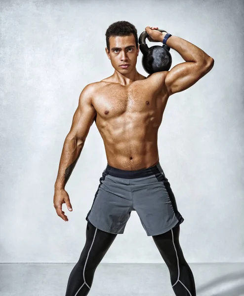 Sporty young man working out with a kettlebell. Photo of muscular man with perfect physique on grey background. Strength and motivation