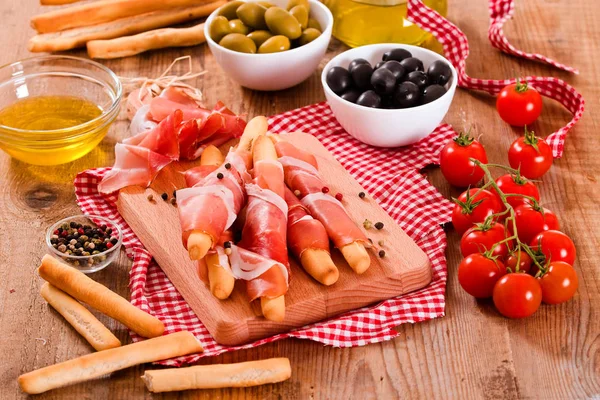 Grissini breadsticks with ham and olives on wooden table. — Stock Photo, Image