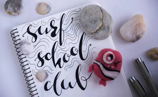 Surf school club black hand written ink in the sketchbook. Against the background stones sea shells and small fish handmade wool