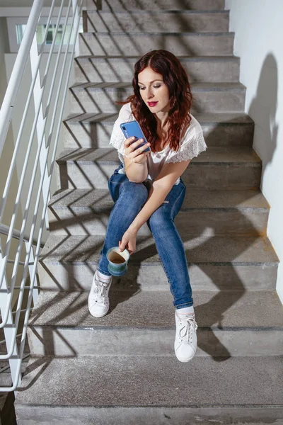 Rotschopf sitzt auf der Treppe und betrachtet Nachrichten auf ihrem Smartphone — Stockfoto