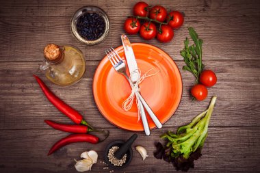 Taze domates, biber ve diğer baharatlar ve otlar ahşap masa merkezinde modern turuncu plaka çevresinde. Üstten Görünüm. Çatal ve bıçak plaka üzerinde. Yatay.