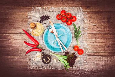 Taze domates, biber ve diğer baharatlar ve otlar ahşap masa ve bez peçete merkezinde modern turkuaz plaka çevresinde. Çatal ve bıçak plaka üzerinde bağlı. Üstten Görünüm. Yatay.