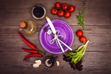 Taze domates, biber ve diğer baharatlar ve otlar ahşap masa merkezinde modern mor plaka çevresinde. Üstten Görünüm. Çatal ve bıçak plaka üzerinde. Yatay.