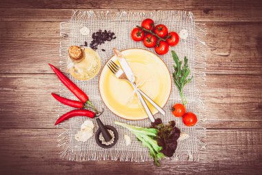 Taze domates, biber ve diğer baharatlar ve otlar ahşap masa ve bez peçete merkezinde modern sarı plaka çevresinde. Çatal ve bıçak plaka üzerinde bağlı. Üstten Görünüm. Yatay.