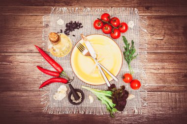 Taze domates, biber ve diğer baharatlar ve otlar ahşap masa ve bez peçete merkezinde modern sarı plaka çevresinde. Çatal ve bıçak plaka üzerinde bağlı. Üstten Görünüm. Yatay.