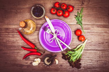 Taze domates, biber ve diğer baharatlar ve otlar ahşap masa merkezinde modern mor plaka çevresinde. Üstten Görünüm. Çatal ve bıçak plaka üzerinde. Yatay.