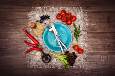 Taze domates, biber ve diğer baharatlar ve otlar ahşap masa ve bez peçete merkezinde modern turkuaz plaka çevresinde. Çatal ve bıçak plaka üzerinde bağlı. Üstten Görünüm. Yatay.