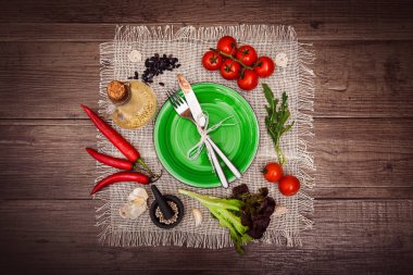 Taze domates, biber ve diğer baharatlar ve otlar ahşap masa ve bez peçete merkezinde modern yeşil plaka çevresinde. Çatal ve bıçak plaka üzerinde bağlı. Üstten Görünüm. Yatay.