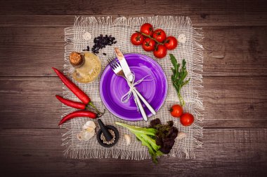 Taze domates, biber ve diğer baharatlar ve otlar ahşap masa ve bez peçete merkezinde modern mor plaka çevresinde. Çatal ve bıçak plaka üzerinde bağlı. Üstten Görünüm. Yatay.